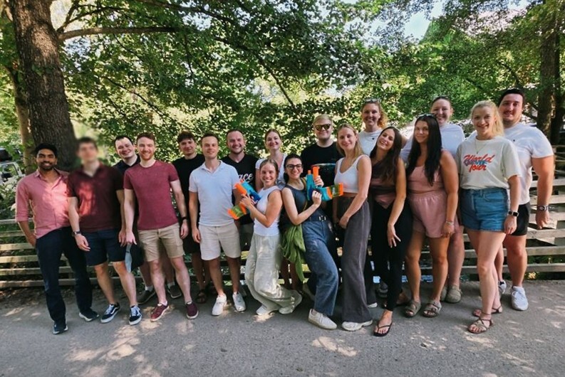 Gruppenfoto des summ.ai Teams beim Offsite 2026. Rund 20 Teammitglieder stehen lächelnd im Freien unter grünen Bäumen, zwei Kolleginnen in der Mitte halten Wasserpistolen.