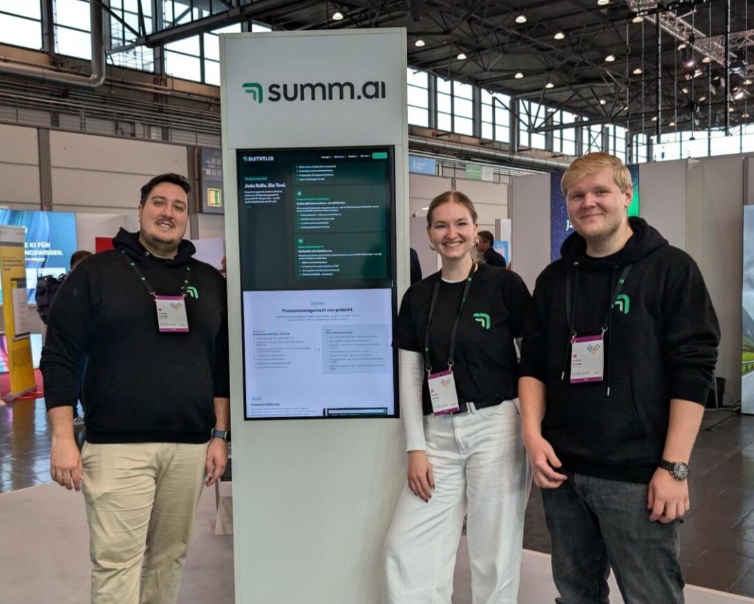 Fabian Langer, Emilia Zaiser und Julian Novak (von links) von summ.ai stehen lächelnd neben einem Messestand mit summ.ai Bildschirm auf dem KGSt-Forum 2026.