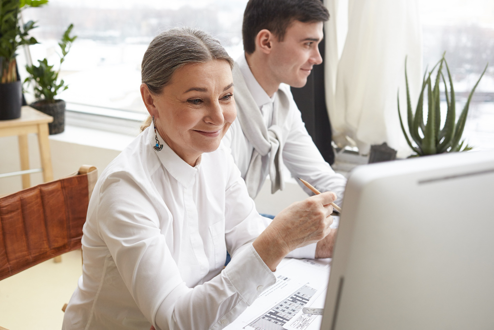 Eine ältere Frau mit grauen Haaren sitzt lächelnd am Schreibtisch vor einem Computer, ein jüngerer Kollege sitzt neben ihr in einem hellen Büro.