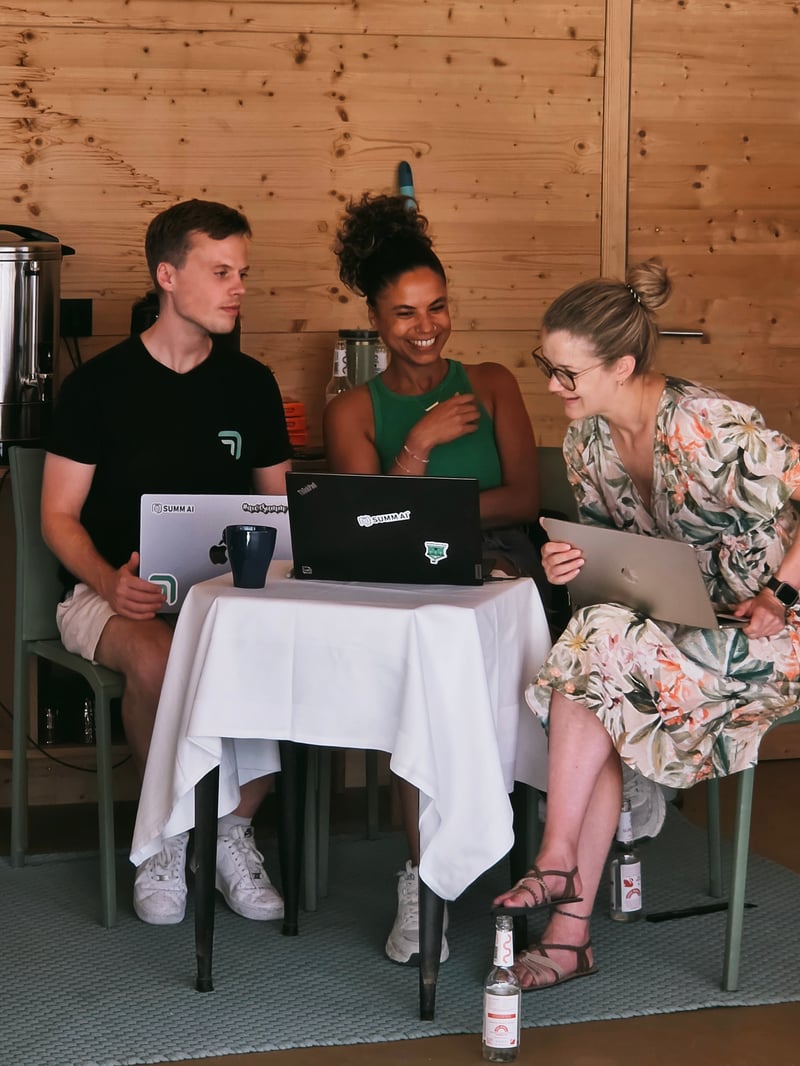 : Drei Teammitglieder von summ.ai sitzen beim Offsite 2026 mit Laptops an einem kleinen Tisch und besprechen lachend etwas.