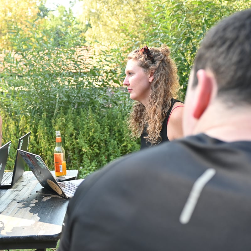 Eine Teammitglied von summ.ai hört beim Offsite 2026 an einem Tisch im Grünen einer Diskussion zu, im Vordergrund Laptops und ein Kollege von hinten.