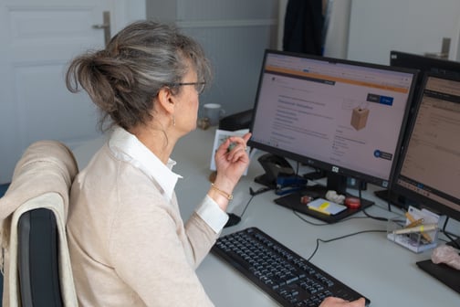 Eine Frau mit grauen Haaren sitzt an einem Schreibtisch mit zwei Bildschirmen und liest konzentriert einen Text auf einer Website.