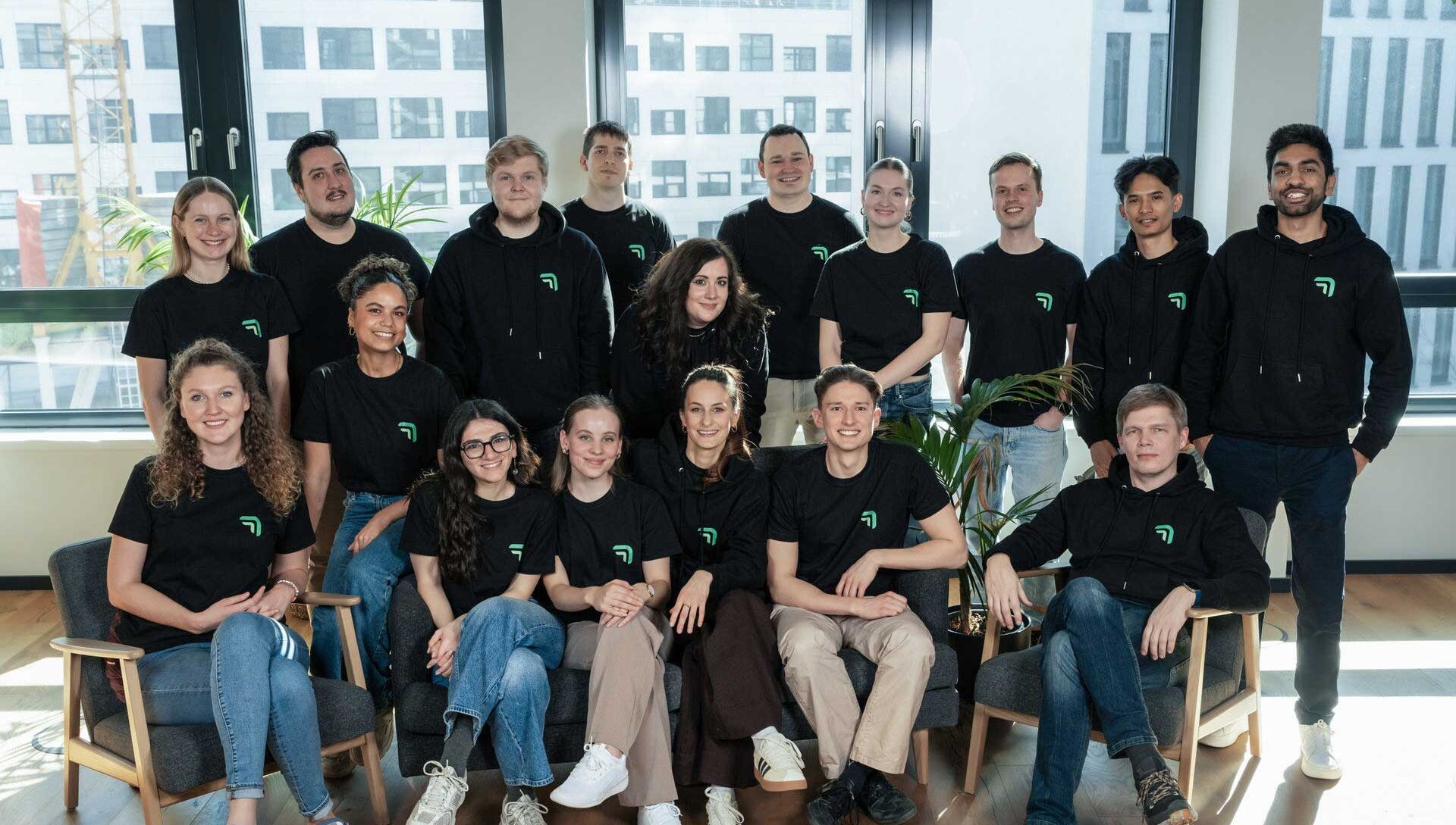Gruppenfoto des summ.ai Teams in schwarzen Shirts und Hoodies mit Logo. Ein Teil sitzt vorne auf Sesseln und einem Sofa, der Rest steht dahinter vor großen Bürofenstern.