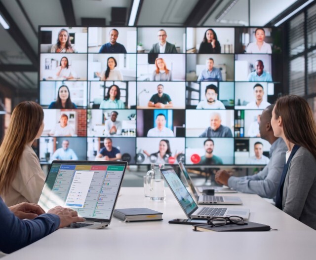 Mehrere Personen sitzen mit Laptops an einem Konferenztisch und schauen auf eine große Videowand, auf der viele Teilnehmende per Videocall zugeschaltet sind. Symbolbild für den Community Stammtisch von summ.ai als Webinar.