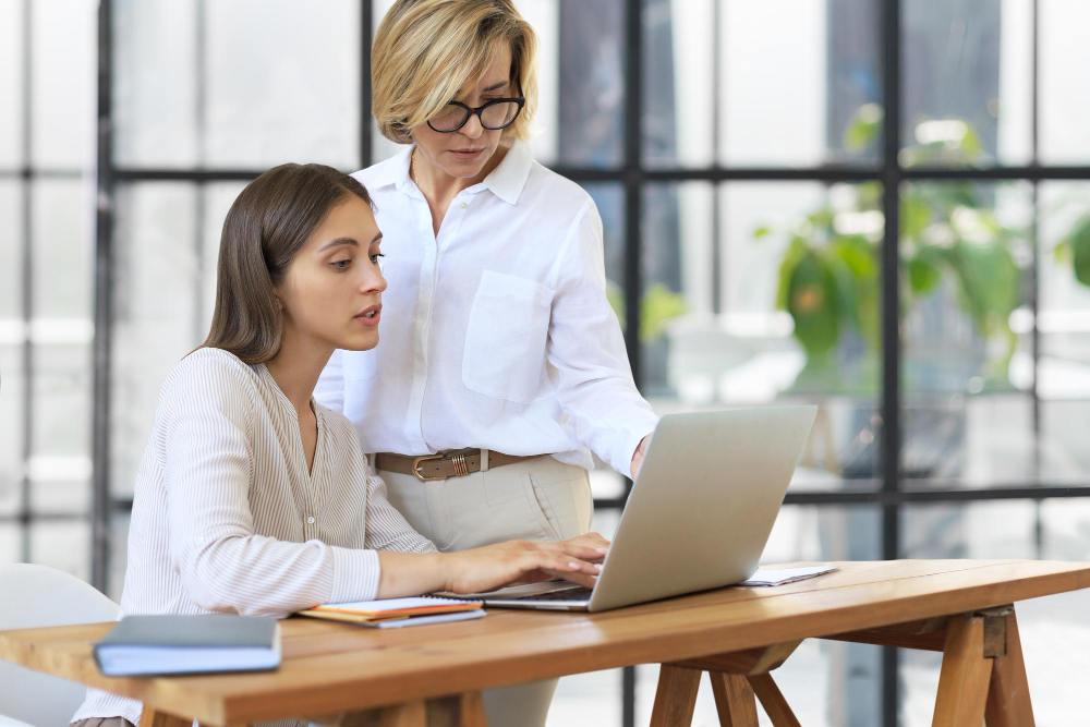 Eine junge Frau arbeitet an einem Laptop an einem Holztisch, eine Kollegin steht neben ihr und schaut auf den Bildschirm.