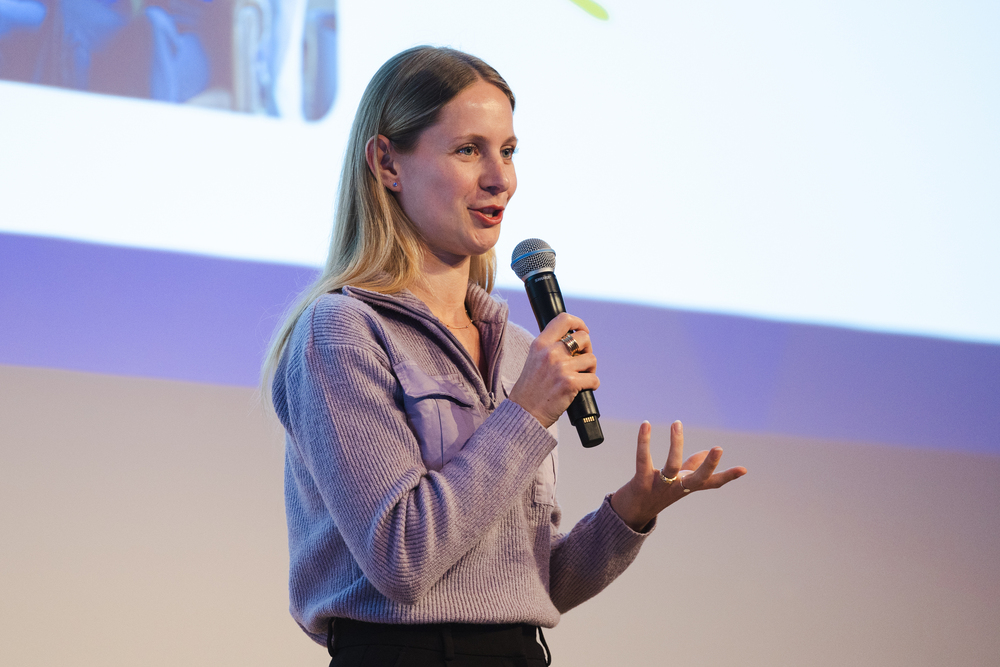 Vanessa König, CRO von summ.ai, spricht mit Mikrofon auf einer Bühne und unterstreicht ihre Worte mit einer Handbewegung, hinter ihr eine Projektionsfläche.