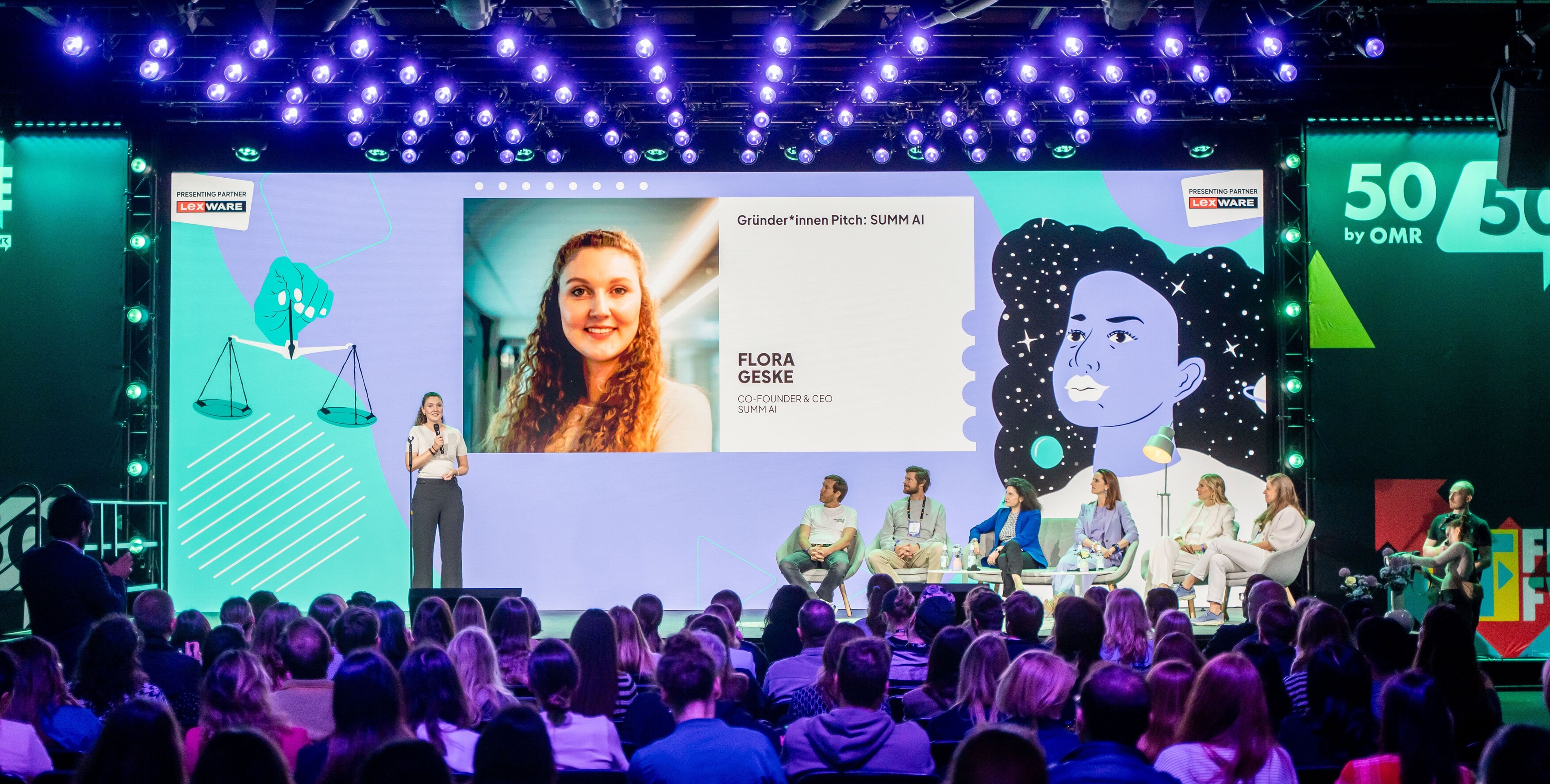 Flora Geske, CEO von summ.ai, pitcht summ.ai mit Mikrofon auf der Bühne von 50/50 by OMR vor vollem Saal. Hinter ihr zeigt eine große Leinwand ihr Foto und ihren Namen, rechts auf der Bühne sitzt die Jury.