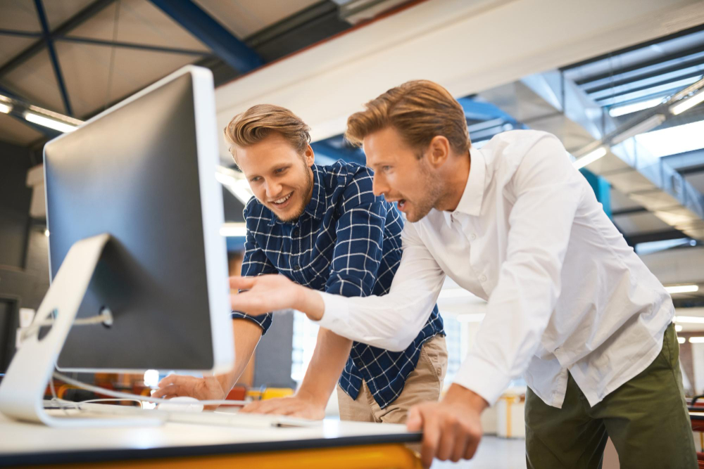 Zwei Männer stehen in einem modernen Büro vor einem Computerbildschirm, einer zeigt lächelnd auf den Monitor.