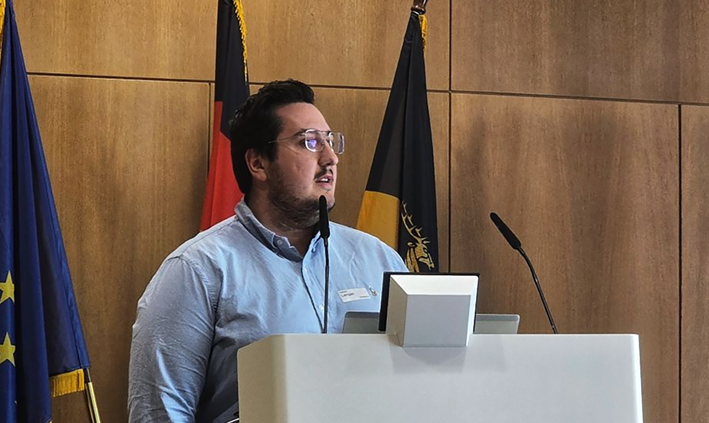 Fabian Langer von summ.ai hält einen Vortrag an einem Rednerpult, hinter ihm stehen die Flaggen der EU, Deutschlands und eines Bundeslandes.
